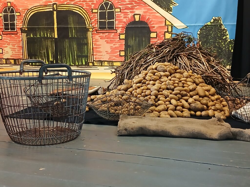 Ein Haufen Kartoffeln mit trockenem Kartoffelkraut liegt neben einem Jutesack und zwei Drahtkörben auf einem Holzboden vor einer gemalten Bühnenkulisse mit rotem Backsteinhof und blauem Himmel.
