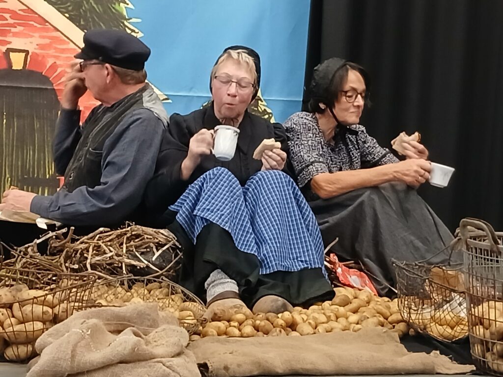 Drei Darsteller in historischer ländlicher Kleidung sitzen bei einer Theateraufführung der Platt AG der Grafschaft Bentheim zwischen Kartoffeln, Drahtkörben und Jutesäcken auf der Bühne und machen mit Brot und Kaffeetassen eine Pause bei der Kartoffelernte.