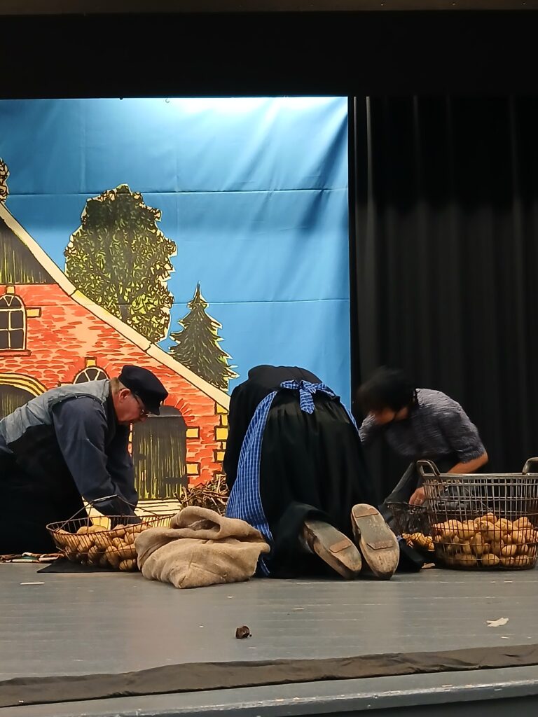 Drei Darsteller in ländlicher Kleidung knien auf einer Theaterbühne zwischen Drahtkörben voller Kartoffeln und Jutesäcken vor einer gemalten Kulisse mit rotem Backsteinhaus und Bäumen, eine Frau mit Holzschuhen und blauer Schürze in der Mitte.