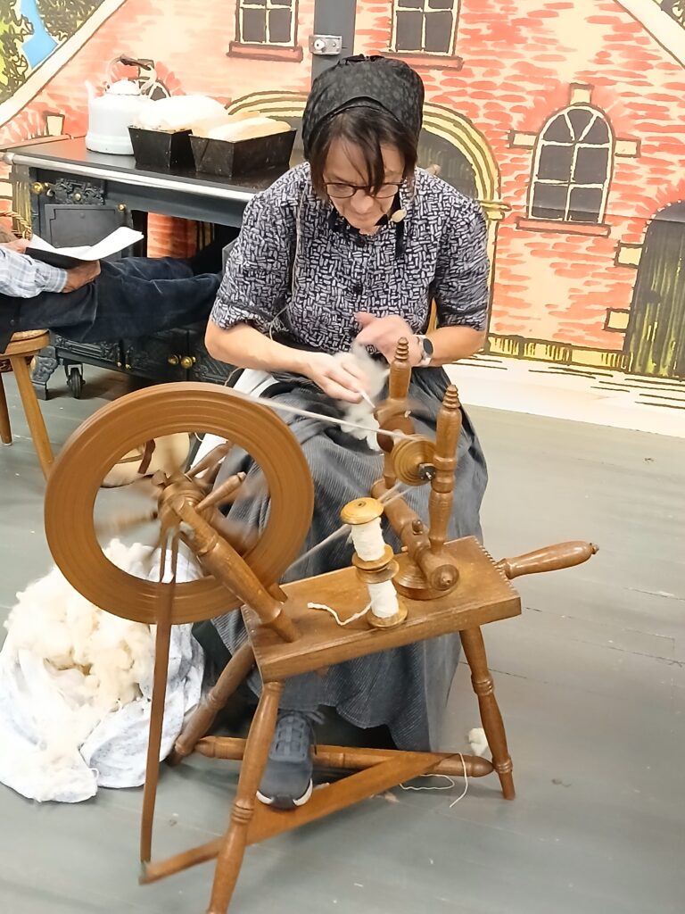Eine Frau in historischer Tracht mit Haube und Brille spinnt an einem hölzernen Spinnrad Wolle zu Garn, vor einer bemalten Kulisse mit Backsteinhaus und einem alten Küchenherd mit Kessel und Backformen.