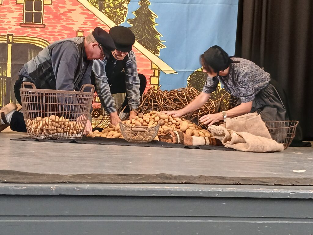 Drei Darsteller in traditioneller ländlicher Kleidung knien auf einer Bühne vor einer gemalten Dorfkulisse und sortieren Kartoffeln in Drahtkörbe – eine Szene aus einer plattdeutschen Theateraufführung.