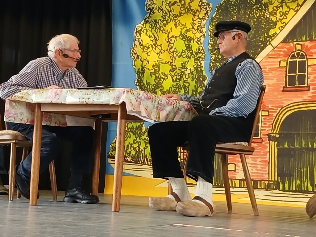 Zwei ältere Schauspieler mit Headset-Mikrofonen sitzen bei einer plattdeutschen Theateraufführung an einem Tisch mit Blümchendecke vor einer bunt gemalten Kulisse mit Backsteinhaus und Bäumen, einer von ihnen in Schiebermütze, Weste und Holzschuhen.