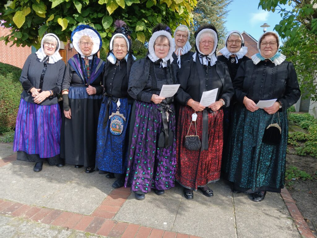 **Alt-Text:**„Acht ältere Frauen in traditioneller Grafschafter Tracht mit weißen Rüschenhauben, schwarzen Jacken und bunten Seidenröcken stehen lächelnd vor einem Baum und einem Backsteingebäude, drei von ihnen halten Textblätter in den Händen.**Hinweis:** Du hast nur ein Bild hochgeladen, deshalb habe ich nur dafür einen Alt-Text erstellt. Wenn du weitere Bilder schickst, schreibe ich gern auch für diese passende Beschreibungen. Falls du weißt, um welchen Anlass es sich handelt (z. B. ein plattdeutscher Gottesdienst oder eine Lesung), kann ich den Alt-Text entsprechend ergänzen. Das habe ich hier bewusst nicht getan, weil es auf dem Bild nicht eindeutig erkennbar ist.