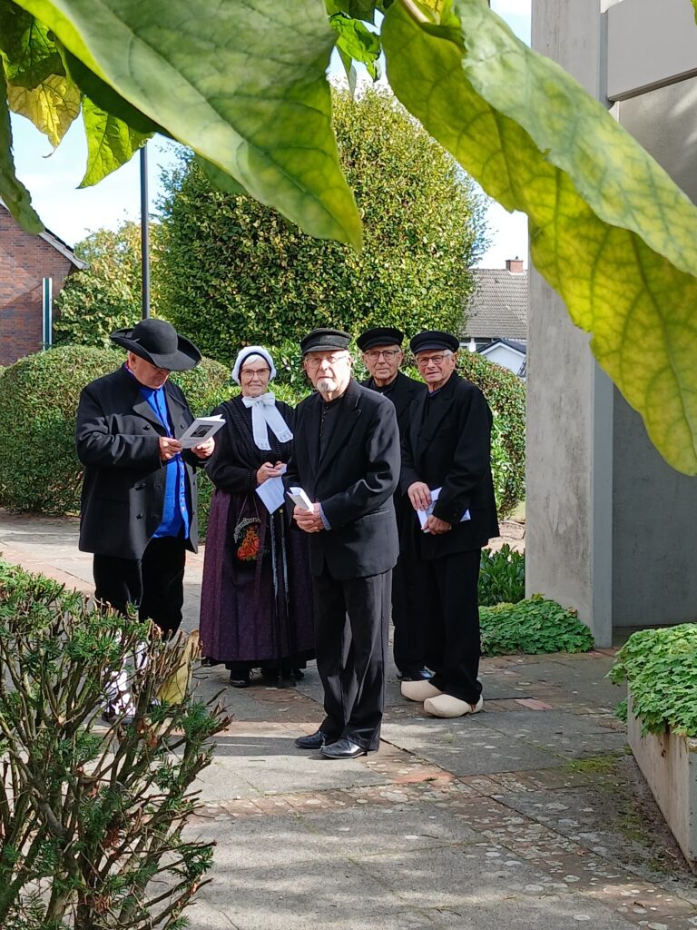**Alt-Text:**„Fünf Personen in traditioneller, dunkler Tracht, darunter eine Frau mit weißer Haube und vier Männer mit Hüten oder Schirmmützen, stehen mit kleinen Heften in den Händen auf einem Weg zwischen Hecken und großen Blättern; einer der Männer trägt hellgelbe Holzschuhe.**Hinweis:** Sie schreiben „für jedes Bild, ich sehe aber nur dieses eine Bild. Wenn Sie weitere Bilder hochladen, erstelle ich gern auch dafür passende Alt-Texte. Falls Sie den Anlass kennen, etwa einen plattdeutschen Gottesdienst, eine Lesung oder einen Auftritt der Platt AG, kann ich den Alt-Text entsprechend ergänzen.