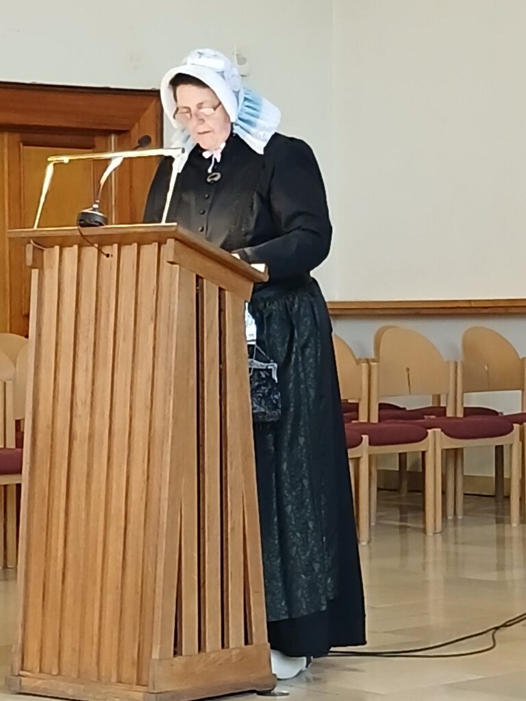 **Alt-Text:**
Eine Frau in historischer Tracht mit schwarzem Kleid, dunkler Schürze und weißer Haube mit hellblauer Rüsche liest an einem hölzernen Rednerpult mit Mikrofon einen Text vor, im Hintergrund sind Stuhlreihen in einem hellen Raum zu sehen.Du hast nur ein Bild hochgeladen. Wenn noch weitere Bilder folgen sollen, schick sie gern, dann erstelle ich für jedes einen eigenen Alt-Text. Wenn du mir mehr Kontext gibst, etwa den Anlass, die Veranstaltung oder den Namen der Person, kann ich den Text noch besser an deine Webseite anpassen. Zum Beispiel: „Eine Frau in historischer Tracht liest bei einer Veranstaltung der Platt AG der Grafschaft Bentheim einen plattdeutschen Text vor.“