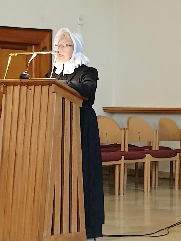 **Alt-Text:** Eine ältere Frau in historischer dunkler Kleidung mit weißer Haube und Brille steht an einem hölzernen Rednerpult mit Mikrofonen und spricht in einem hellen Raum mit gepolsterten Stühlen im Hintergrund.Ich habe nur dieses eine Bild erhalten. Wenn du die weiteren Bilder der Webseite hochlädst, erstelle ich gern für jedes einen passenden Alt-Text. Falls du mir den Anlass nennst, etwa eine Lesung auf Plattdeutsch oder einen Vortrag des Vereins, kann ich den Text noch genauer auf den Zusammenhang der Webseite abstimmen.