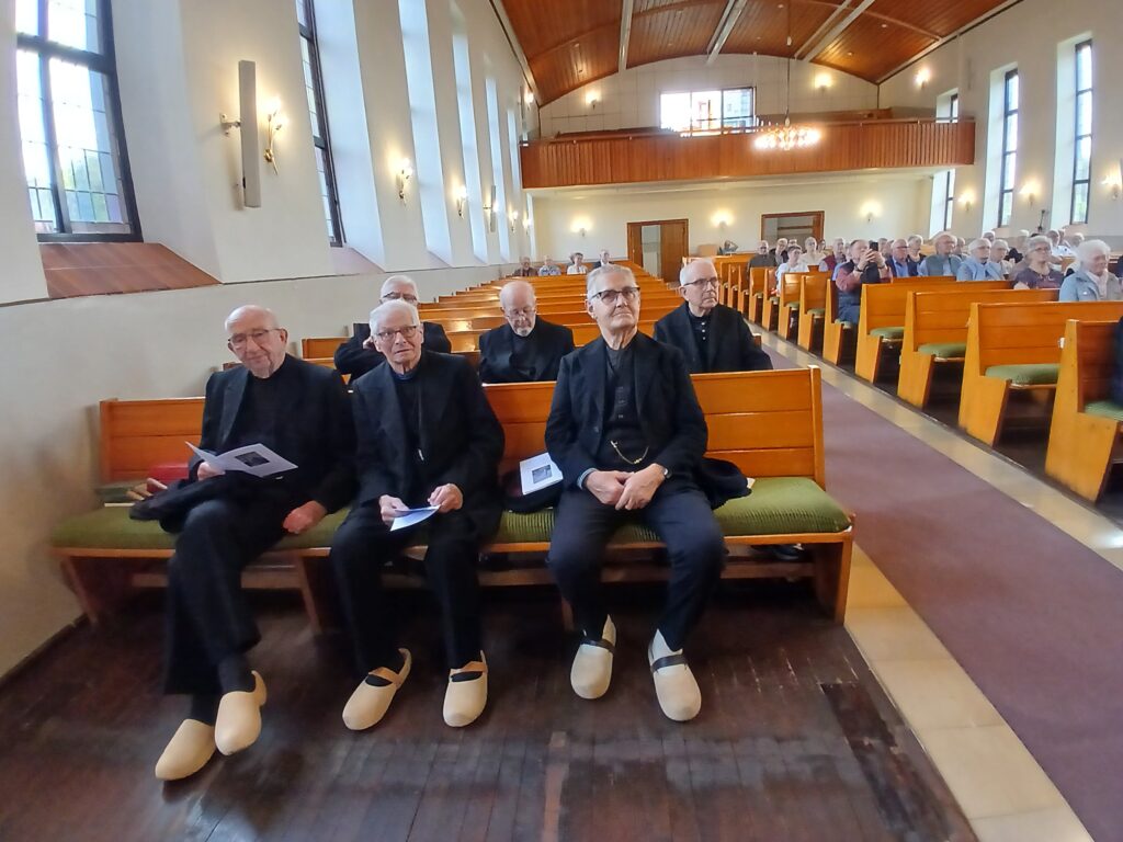 **Alt-Text:** Mehrere ältere Männer in traditioneller dunkler Kleidung und mit hellen Holzschuhen sitzen auf einer Kirchenbank, während sich hinter ihnen der Kirchenraum mit weiteren Besucherinnen und Besuchern füllt.Du hast nur ein Bild hochgeladen. Wenn du weitere Bilder für die Webseite der Platt AG der Grafschaft Bentheim hast, schicke sie mir gern, dann erstelle ich für jedes einen passenden Alt-Text.Falls du bei diesem Bild lieber den Anlass nennen möchtest, etwa einen plattdeutschen Gottesdienst, kann ich den Text entsprechend anpassen.
