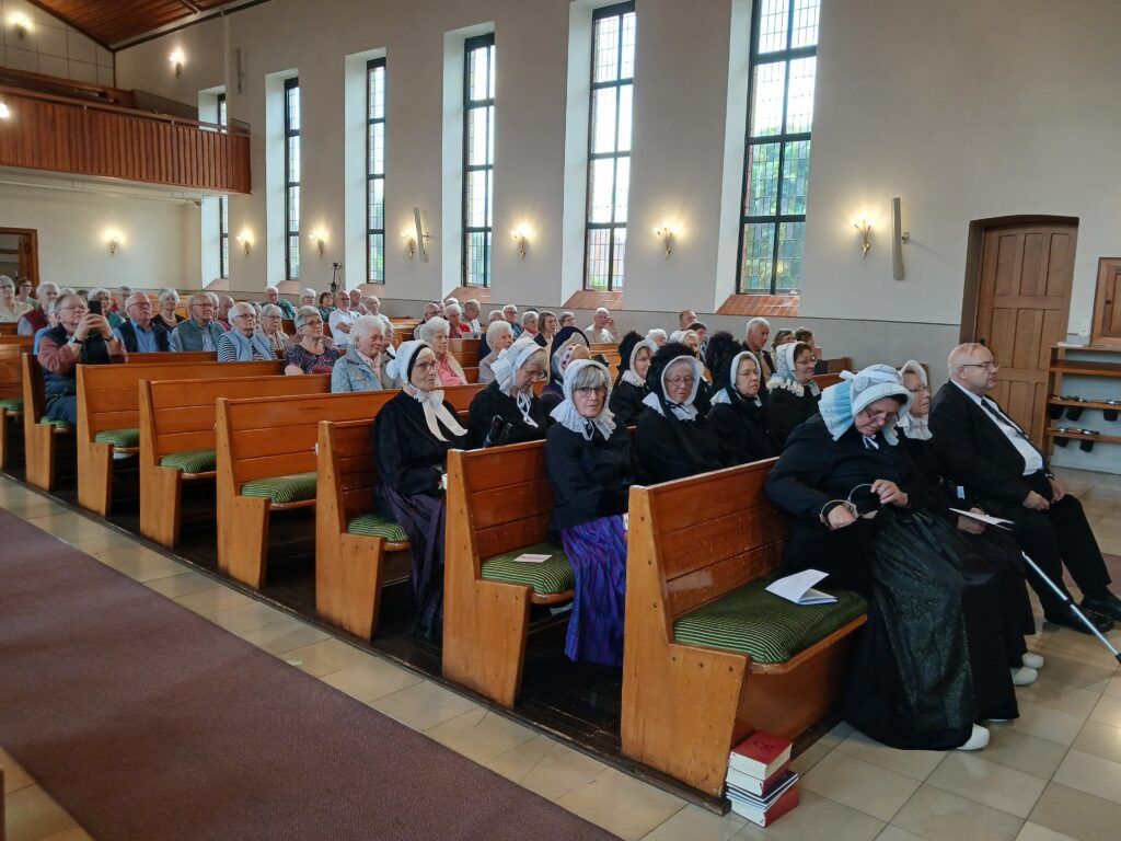 **Alt-Text:** Im Kirchenraum sitzen zahlreiche ältere Besucherinnen und Besucher in Holzbänken, darunter mehrere Frauen in schwarzer, traditioneller Tracht mit weißen Spitzenhauben und ein Mann im dunklen Anzug, während durch die hohen Fenster Tageslicht einfällt.Hinweis: Du hast nur ein Bild hochgeladen, deshalb gibt es nur einen Alt-Text. Wenn du weitere Bilder schickst, erstelle ich gern für jedes eine eigene Beschreibung. Wenn du möchtest, kann ich auch den Bezug zur Platt AG ergänzen, zum Beispiel: „Publikum in traditioneller Tracht bei einer plattdeutschen Veranstaltung der Platt AG der Grafschaft Bentheim“. Dafür müsste ich allerdings wissen, worum es bei der Veranstaltung ging.