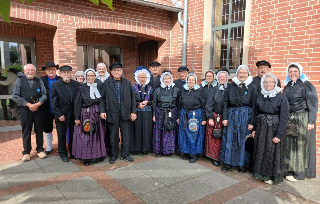 **Alt-Text:**
„Eine Gruppe älterer Frauen und Männer in traditioneller Grafschafter Tracht steht lächelnd vor einem Backsteingebäude; die Frauen tragen dunkle Kleider mit farbigen Schürzen und weiße Hauben, die Männer schwarze Jacken, Westen und Mützen, einige auch Holzschuhe.**Hinweis:** Es wurde nur ein Bild übermittelt, daher habe ich auch nur einen Alt-Text erstellt. Wenn Sie weitere Bilder senden, erstelle ich gern auch für diese passende Beschreibungen. Ich kann den Text auch anpassen, zum Beispiel mit einem Bezug auf „Platt AG der Grafschaft Bentheim“, sofern Sie mir mitteilen, um welche Veranstaltung es sich handelt.