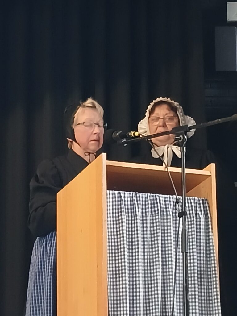 Zwei Frauen in historischer Tracht mit Hauben und karierten Schürzen stehen an einem hölzernen Rednerpult mit Mikrofon vor einem schwarzen Bühnenvorhang und tragen bei einer Veranstaltung der Platt AG der Grafschaft Bentheim auf Plattdeutsch vor.
