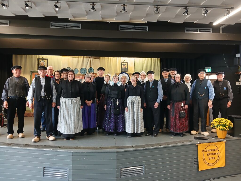 Gruppenfoto von rund 25 Mitgliedern des Groafschupper Plattproater Kring in traditioneller Tracht mit Holzschuhen, Schürzen und Hauben auf einer Bühne vor einer bemalten Kulisse, im Vordergrund ein gelbes Banner mit der Aufschrift „Groafschupper Plattproater Kring – Kreis für Volkskunde und Brauchtum“.