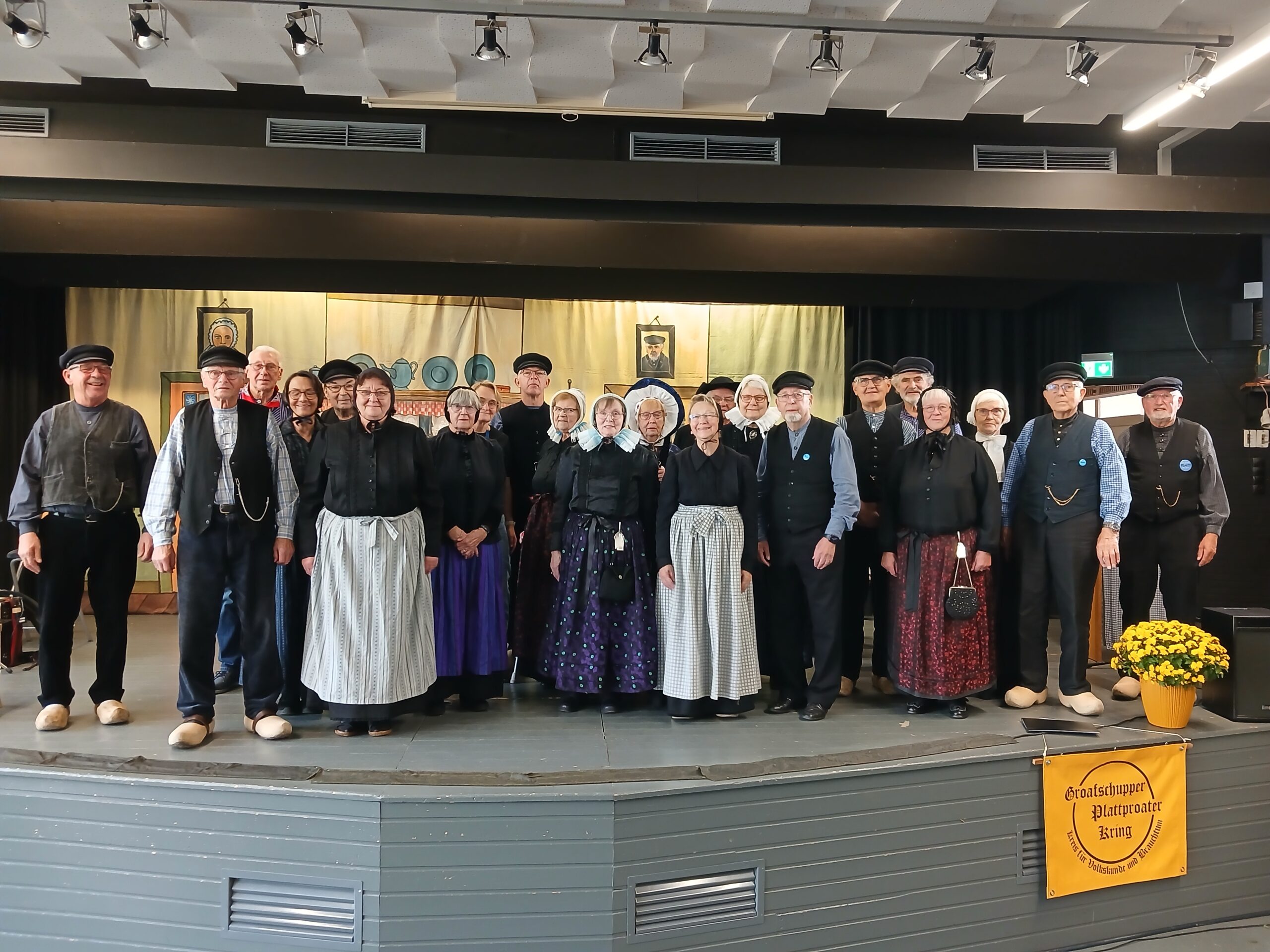 Gruppenfoto von rund 25 Mitgliedern des Groafschupper Plattproater Kring in traditioneller Tracht mit Holzschuhen, Schürzen und Hauben auf einer Bühne vor einer bemalten Kulisse, im Vordergrund ein gelbes Banner mit der Aufschrift „Groafschupper Plattproater Kring – Kreis für Volkskunde und Brauchtum“.