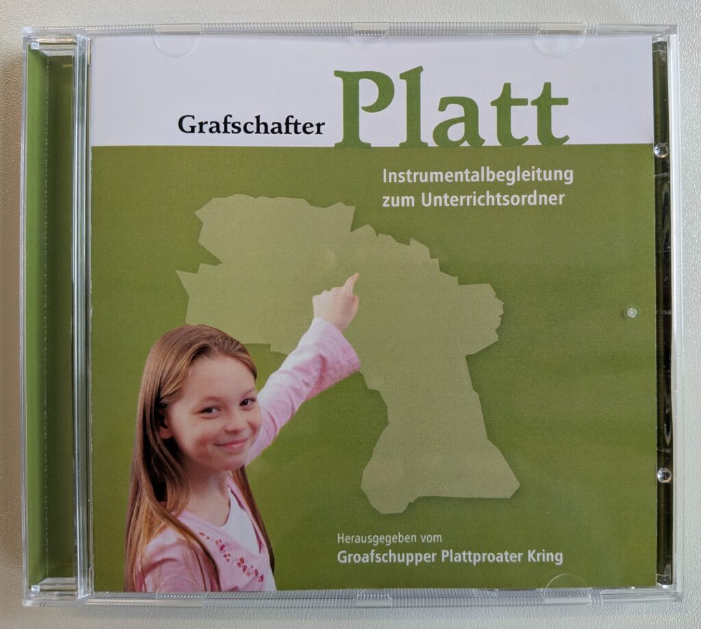 CD-Hülle mit grünem Cover der Instrumentalbegleitung „Grafschafter Platt“ zum Unterrichtsordner, auf dem ein lächelndes Mädchen auf die Umrisskarte der Grafschaft Bentheim zeigt, herausgegeben vom Groafschupper Plattproater Kring.