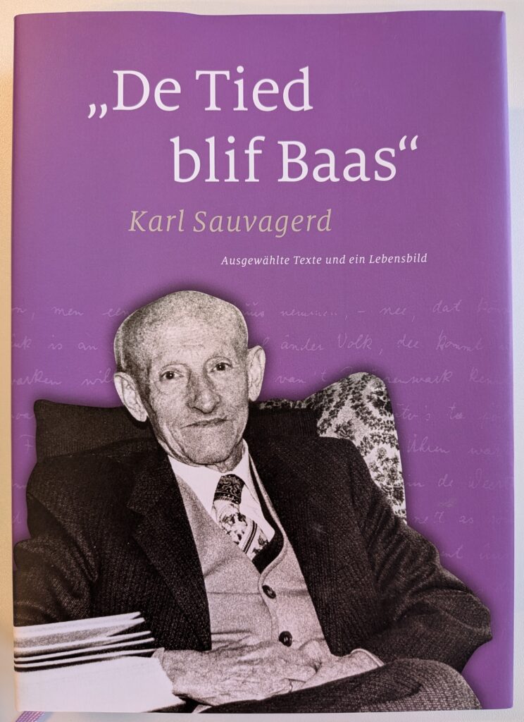 Buchcover in Lila mit dem Titel „De Tied blif Baas“ von Karl Sauvagerd, Untertitel „Ausgewählte Texte und ein Lebensbild“, und einem Schwarz-Weiß-Foto des älteren Autors im Anzug, der lächelnd in einem Sessel sitzt.